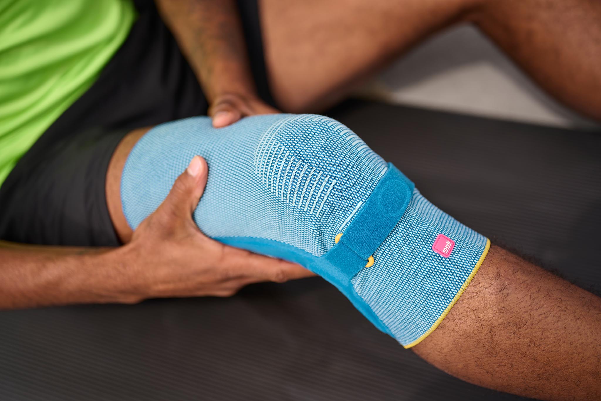 close-up foto van een blauwe Medi kniebrace, gedragen door een Antilliaanse jongen, die speciaal is ontworpen om ondersteuning te bieden bij knieklachten. De persoon houdt met beide handen de kniebrace vast, die om het gebogen been zit.